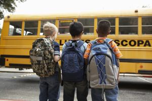 Three boys (8-9) standing with backpacks outdoors near school bus, rear view