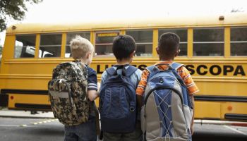 Three boys (8-9) standing with backpacks outdoors near school bus, rear view