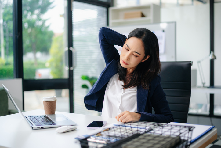 Asian businesswoman feeling neck pain from long office work, stretching at desk with laptop.