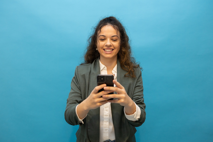 Professional woman smiling, checking smartphone against bright blue backdrop, embodying contemporary workplace energy
