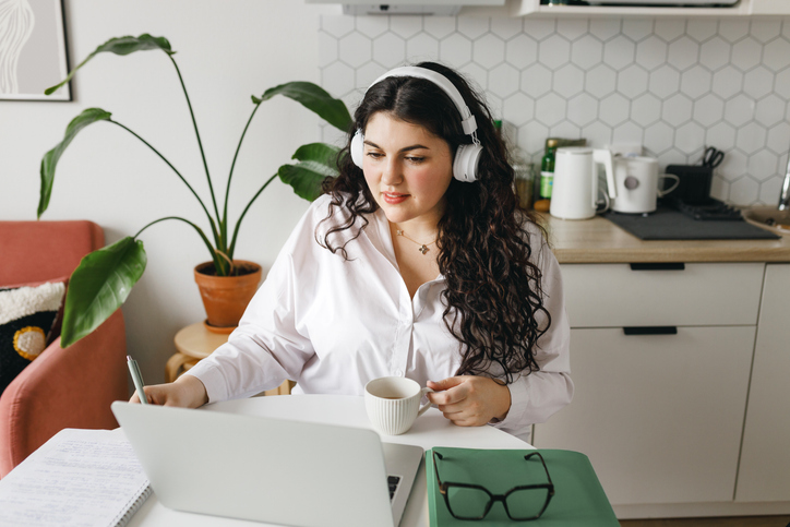 Young adorable woman with curly hair in headphones study online, sitting at kitchen table with coffee in hands, making notes in her copy book after teacher, watching webinar or video tutorial