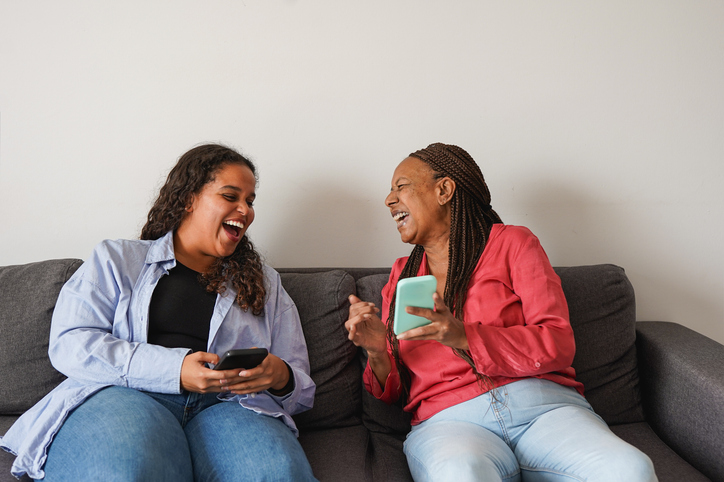 Happy african mother and daughter having fun watching videos on social media with mobile phones at home - Technology, family and lifestyle concept