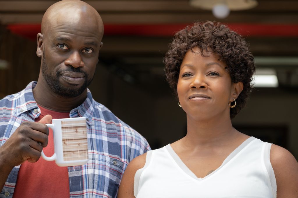 Two adults, a man and a woman, standing together and looking at the camera. The man is wearing a plaid shirt and the woman is wearing a white tank top.