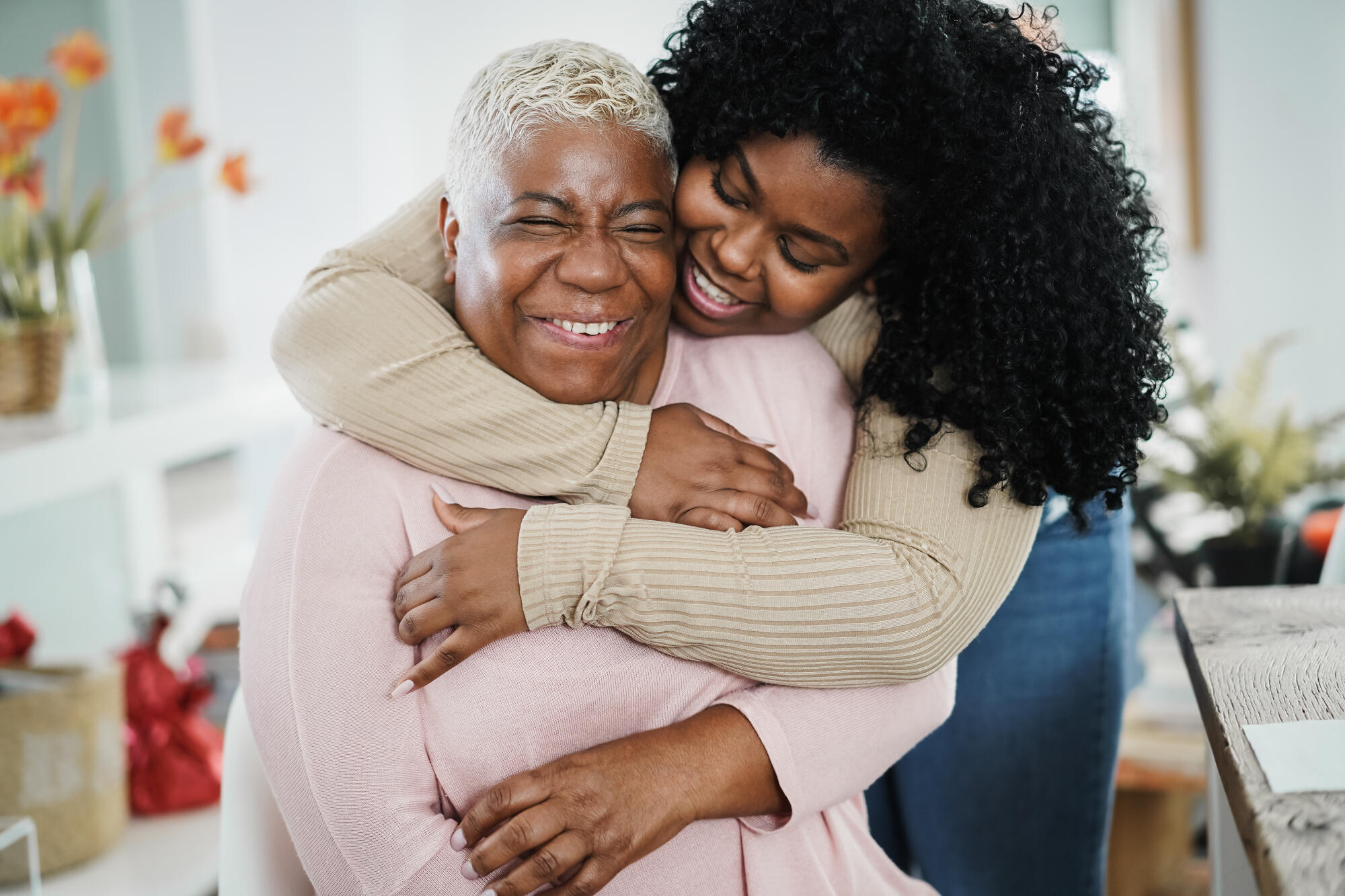 Two women embracing and smiling warmly, one with short gray hair and the other with curly black hair.